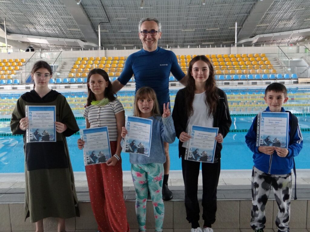 Grup de copii alaturi de instructor, tinand diplomele de absolvire la marginea bazinului, in cadrul scolii Deep Zone Diving.