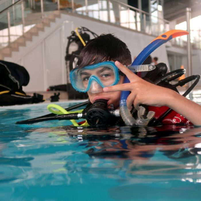 Băiat echipat complet pentru scufundări, în piscină, zâmbește și face semnul victoriei în timpul cursului Try Scuba organizat de Deep Zone Diving.