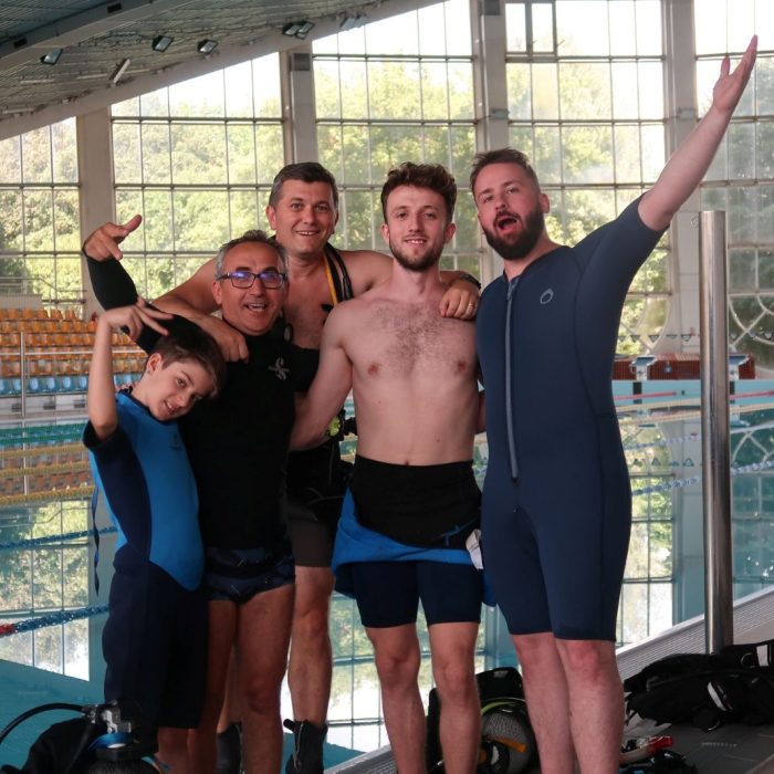 Grup vesel de cursanti si instructori, zambitori si relaxati dupa sesiunea de scuba diving in piscina, la cursul Try Scuba.