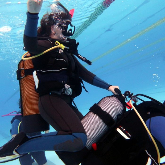 Participanta la cursul Try Scuba sta calare pe spatele unui coleg in piscina, amandoi purtand echipament de scufundare complet.