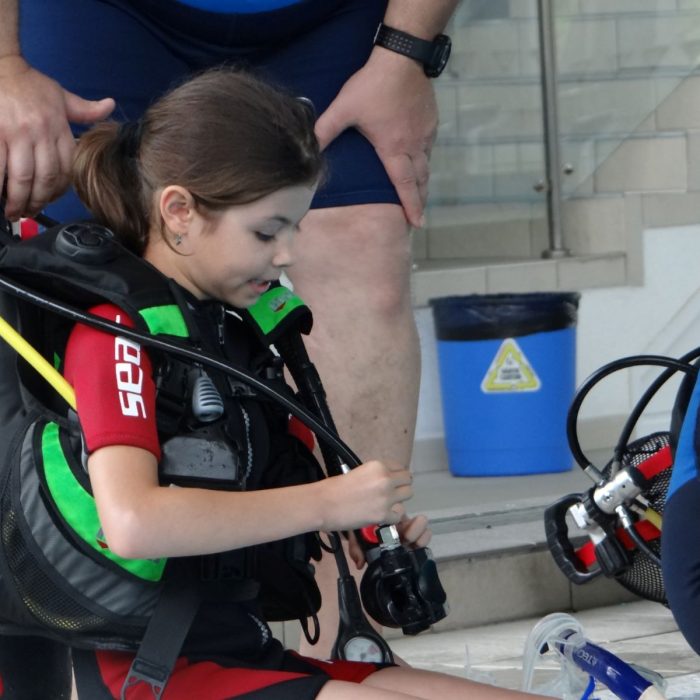 Copil atent echipeaza regulatorul pe butelia de aer, asistat de instructor, in cadrul unei lectii practice de scuba diving pentru incepatori.