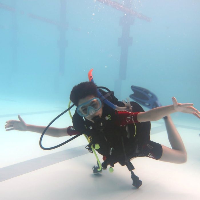Copil plutind relaxat sub apa in piscina, cu brațele intinse si echipament complet, la cursul Try Scuba de initiere in scufundări.