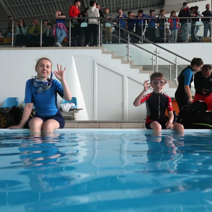 Baiat si fata imbracati in neopren stau pe marginea piscinei si fac semnul OK, in timp ce publicul ii admira din tribuna.