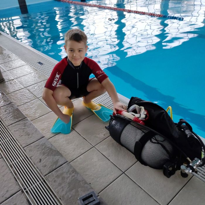 Baiat imbracat in costum de neopren si labe de inot, pregatindu-se pentru o lectie de scufundari langa o piscina interioara.
