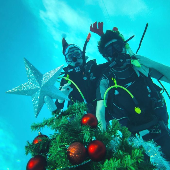 Doi scafandri in costume festive pozeaza langa bradul subacvatic decorat, in timpul unui eveniment organizat de scoala Deep Zone Diving.