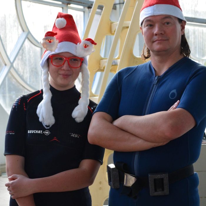 Doua persoane in costume de neopren si caciuli de Mos Craciun zambind in fata camerei, langa bazinul de antrenament Deep Zone Diving.