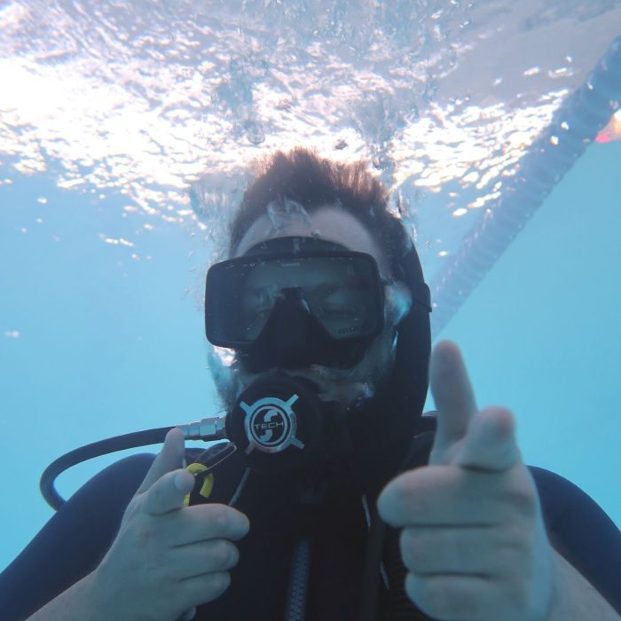 Barbat sub apa, zambitor, face semne cu mainile catre camera in timpul unei sesiuni de scuba diving la cursul Try Scuba.