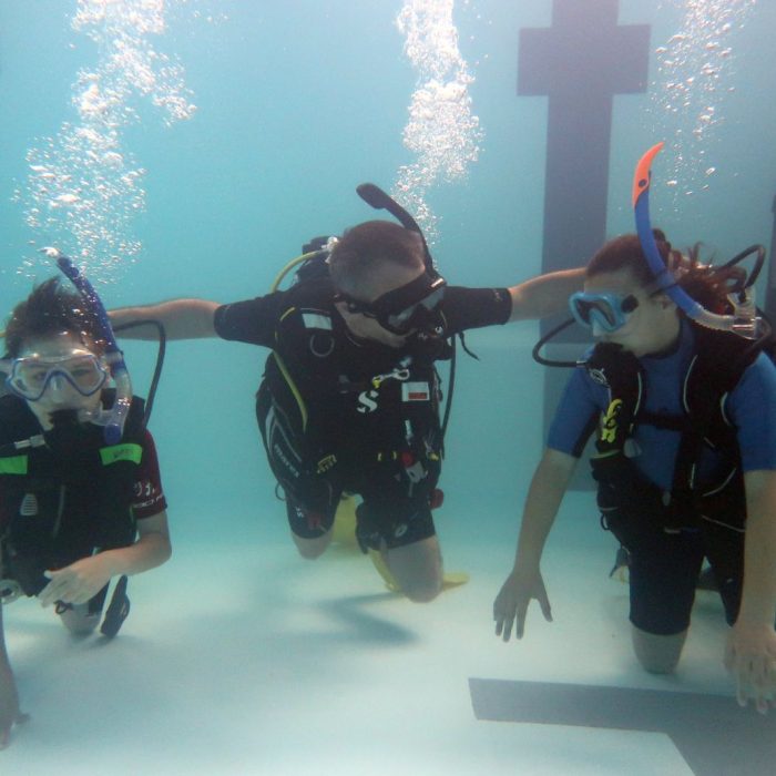 Instructor de scuba diving asistand doi copii sub apa in piscina, oferind suport si siguranta in timpul cursului Try Scuba.