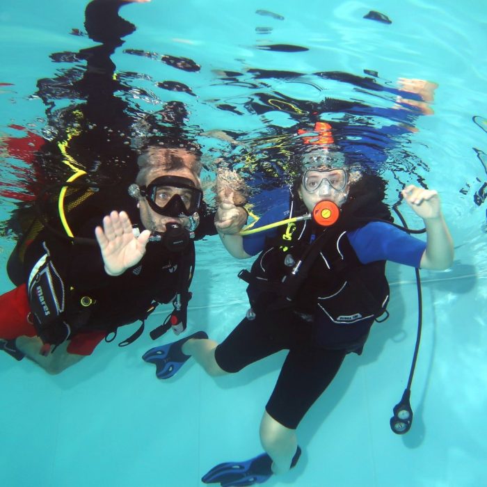 Instructor si cursant echipati complet se tin de mana si zambesc sub apa, in timpul unei sesiuni Try Scuba in piscina.