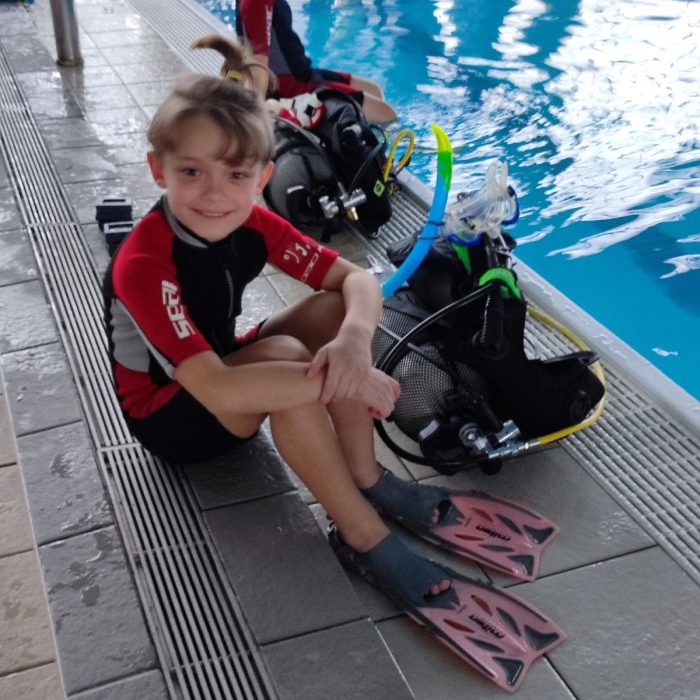 Doi copii stand pe marginea unei piscine interioare, zambind și echipati cu labe, costume si butelii de scufundare.