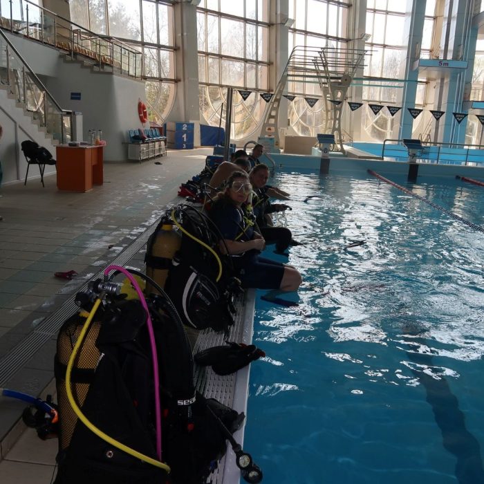 Cursanti echipati complet pentru scufundari, asezati pe marginea unei piscine interioare moderne in timpul unei sesiuni Deep Zone Diving.