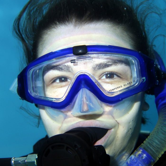 Portret subacvatic al unei cursante cu masca si regulator, surprinsa intr-un moment de calm si curiozitate in timpul unei scufundari.