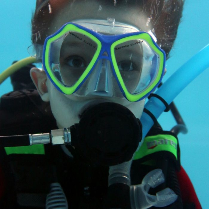 Copil echipat cu masca galben-albastra si regulator in gura, surprins in timpul scufundarii in piscina la cursul Try Scuba.