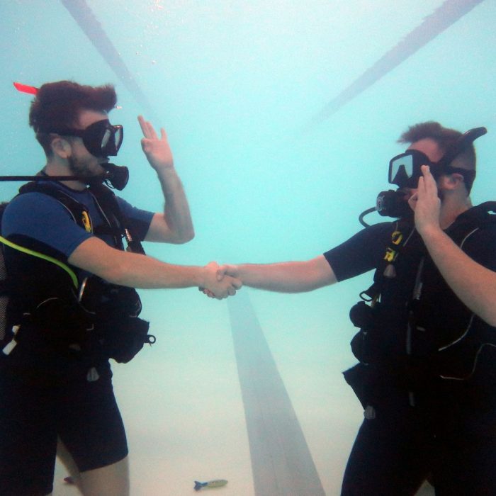Doi cursanti isi strang mana si fac semne de comunicare sub apa, in timpul unui exercitiu la cursul Try Scuba.