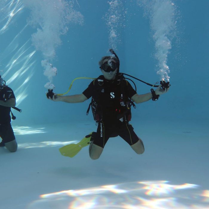 Scafandru efectuează un exercițiu de siguranță cu două regulatoare în mod free flow sub apă, simulând o situație de urgență în cadrul unui antrenament de scufundare în piscină.