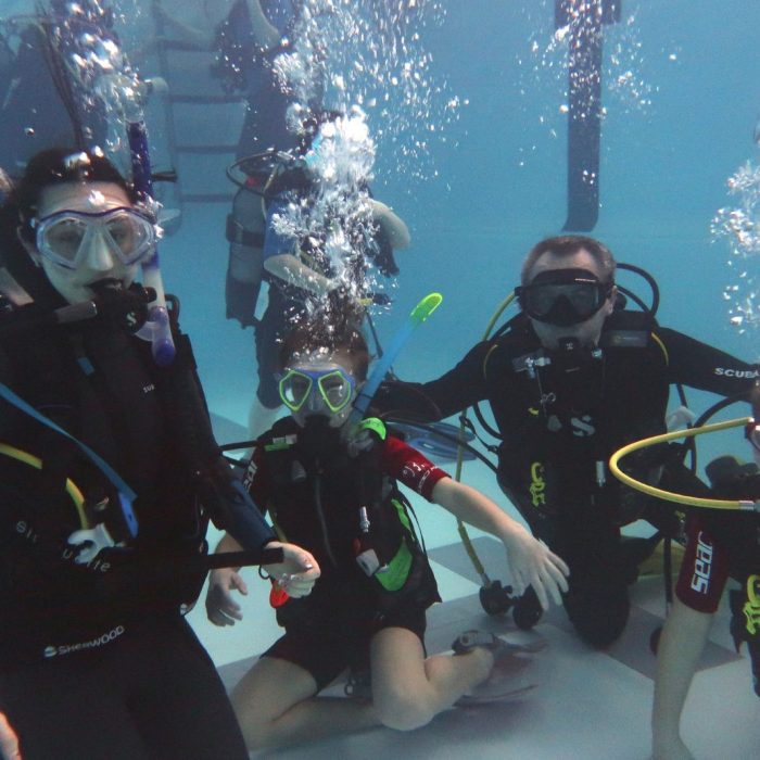 Grup de copii si instructori sub apa, la fundul piscinei, in timpul unei lectii practice de scuba diving pentru incepatori.