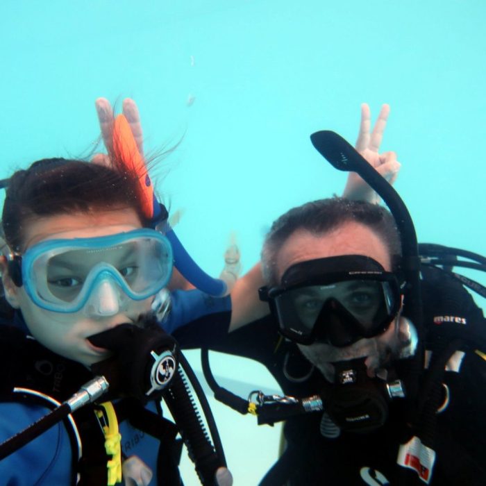 Fetita si instructor scuba sub apa, purtand echipament complet si facand semne jucause in timpul unei sesiuni Try Scuba.