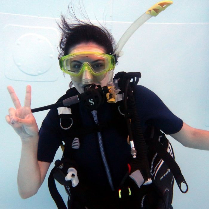 Femeie in echipament complet de scufundare face semnul „peace” in timp ce pluteste sub apa la cursul Try Scuba.