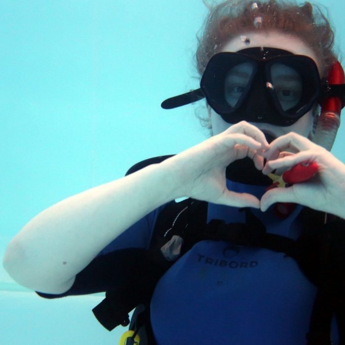 Fata in echipament de scufundare formeaza un semn de inima cu mainile, exprimand entuziasmul si distractia de la cursul Try Scuba.