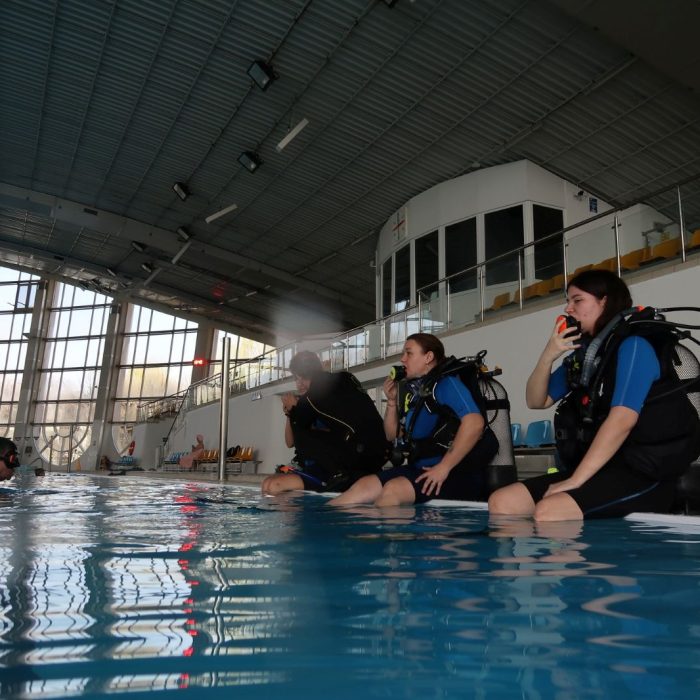 Trei cursante in echipament de scufundare stau pe marginea piscinei si testeaza regulatorul inainte de a intra in apa, sub supravegherea instructorului.