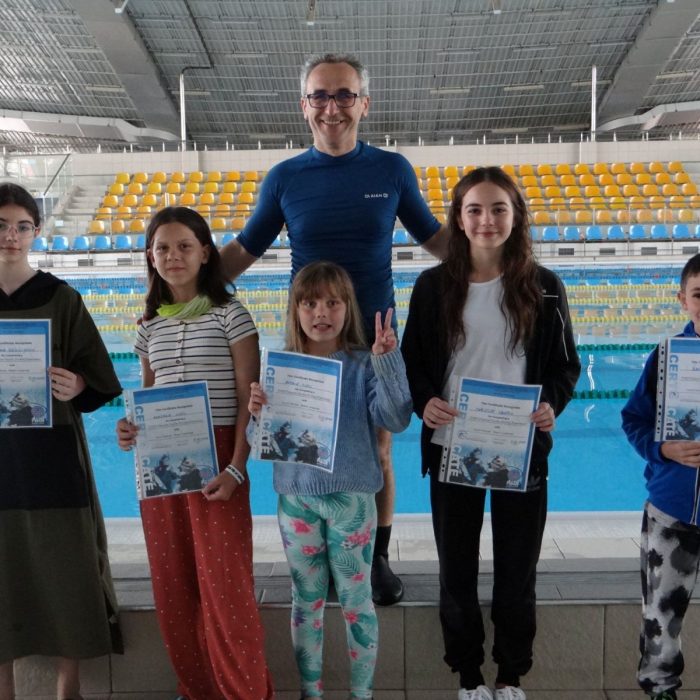 Grup de copii alaturi de instructor, tinand diplomele de absolvire la marginea bazinului, in cadrul scolii Deep Zone Diving.