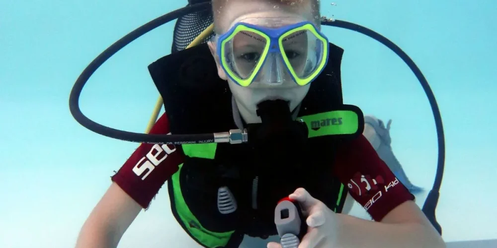 O fotografie subacvatică arată un băiețel, purtând echipament de scufundări, explorând calm fundul unei piscine, pregătit să înceapă cursul de scufundări – Încearcă scuba