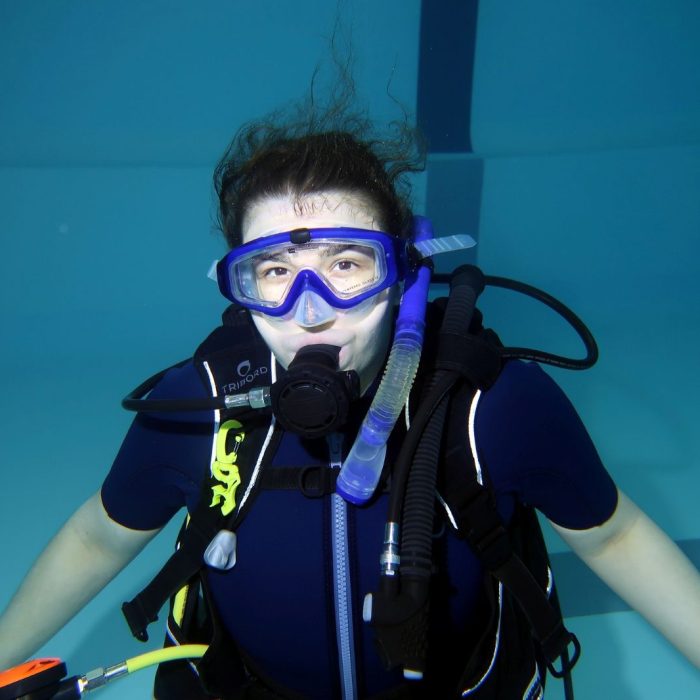 Femeie echipata complet pentru scufundari, zambeste in timp ce pluteste sub apa in cadrul cursului Try Scuba Deep Zone Diving.