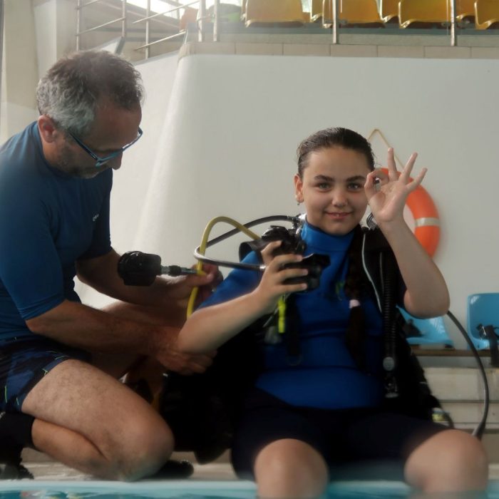 Fata zambitoare face semnul OK in timp ce instructorul o ajuta sa se echipeze pentru prima experienta de scufundare in piscina.